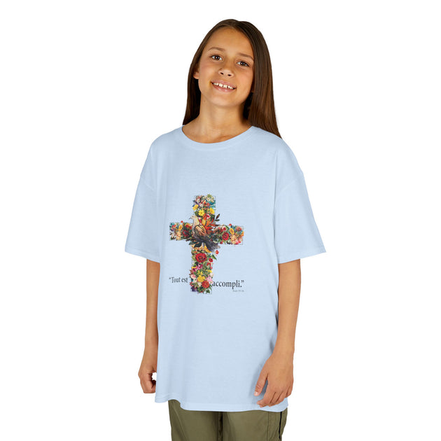 Une enfant porte un t-shirt bleu clair orné d'une croix fleurie et de l'inscription Tout est accompli.