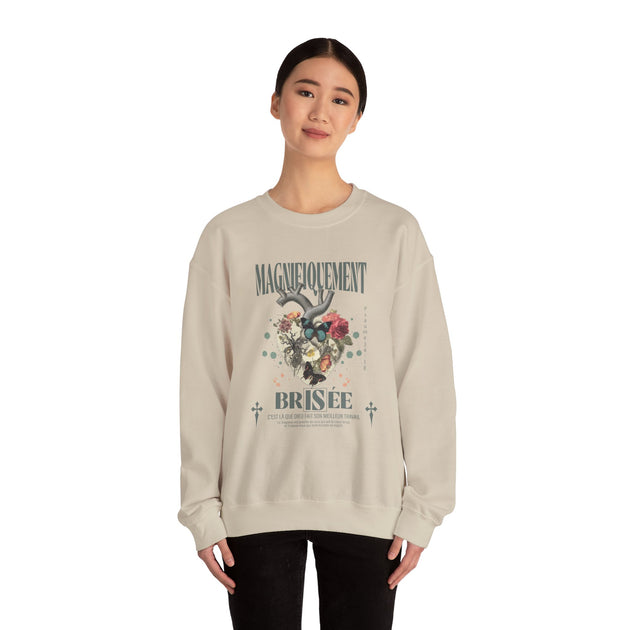 Une femme porte un sweat-shirt beige avec un motif de cœur floral et le texte Magnifiquement Brisée.