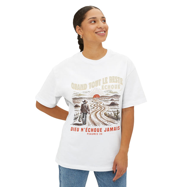 Une femme porte un t-shirt blanc oversize illustré d'un voyageur sur un chemin au soleil levant.