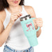 Une femme tient un mug isotherme bleu clair avec un nœud rose et un verset biblique imprimés.