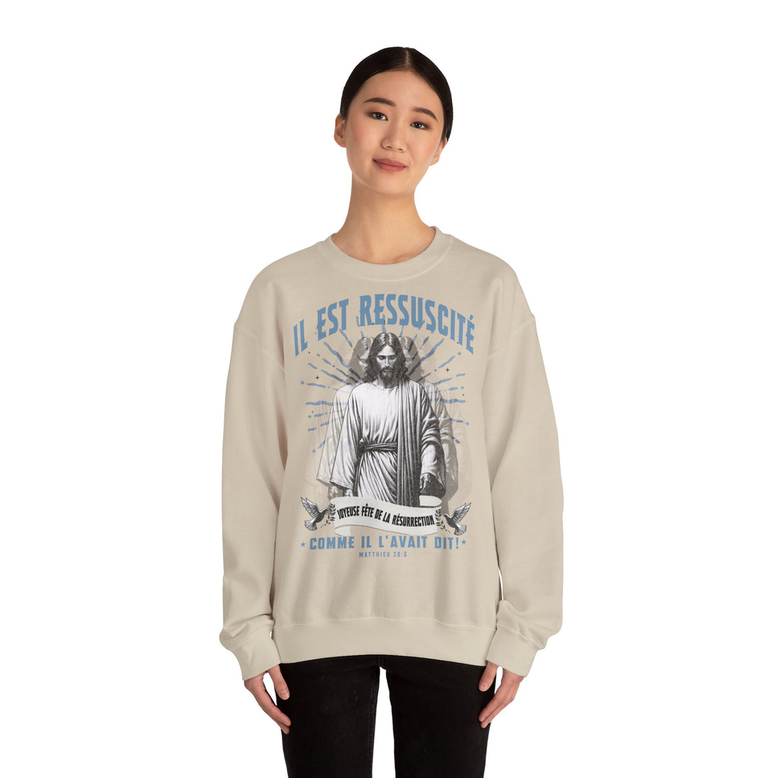 Une femme porte un sweatshirt beige avec une illustration religieuse et le texte Il est ressuscité.