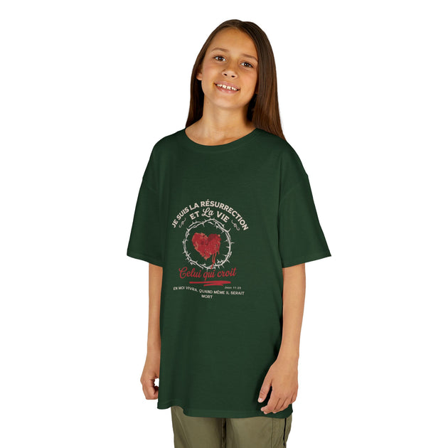 Une enfant porte un t-shirt vert foncé avec un motif religieux représentant un cœur et une couronne.