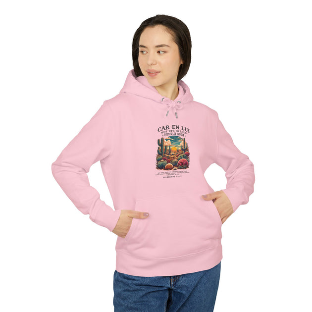 Une femme porte un sweat à capuche rose avec une illustration de cactus et un texte religieux.