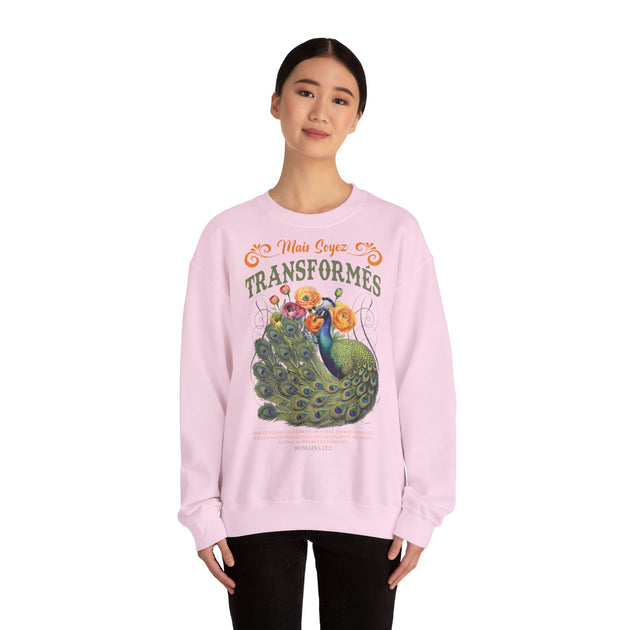 Une femme porte un sweat-shirt rose pâle orné d'un paon, de fleurs et du texte Mais soyez transformés.