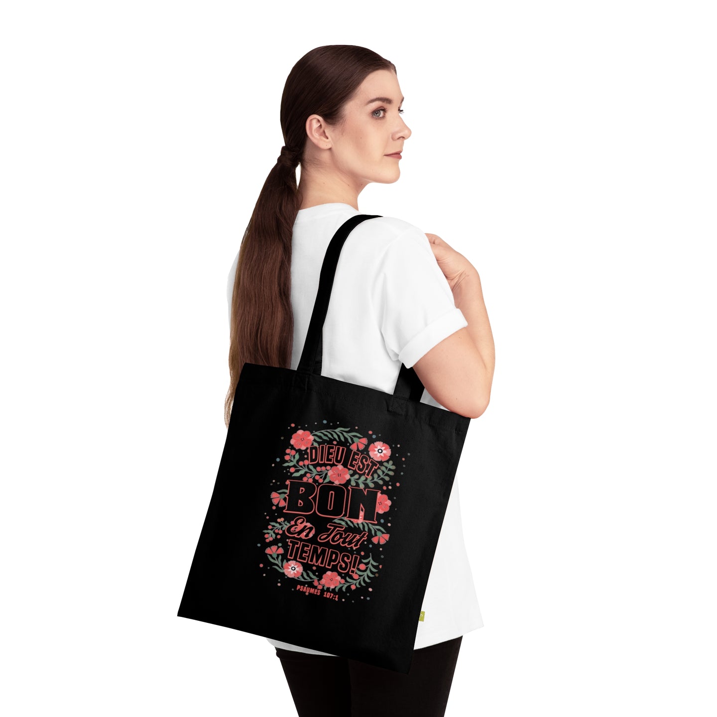 Une femme porte un tote bag noir en coton orné d'un motif floral et du texte Dieu est bon.
