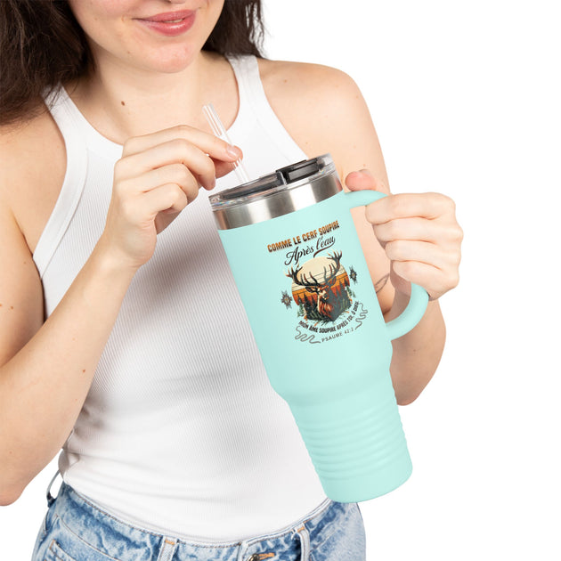 Une femme tient un mug de voyage bleu clair avec une illustration de cerf et un verset biblique.