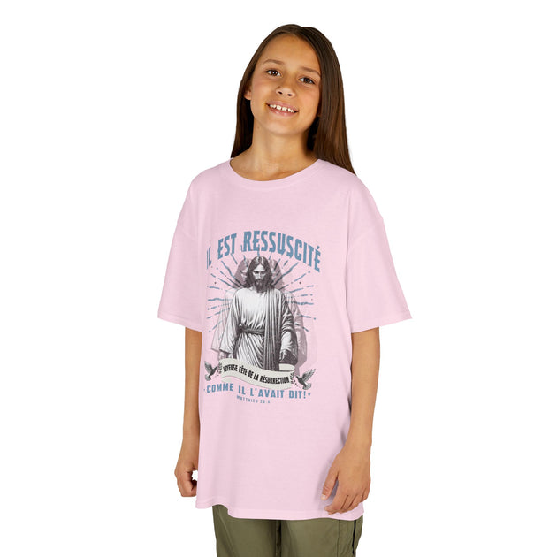 Une jeune fille porte un t-shirt rose clair avec une illustration religieuse et le texte Il est ressuscité.
